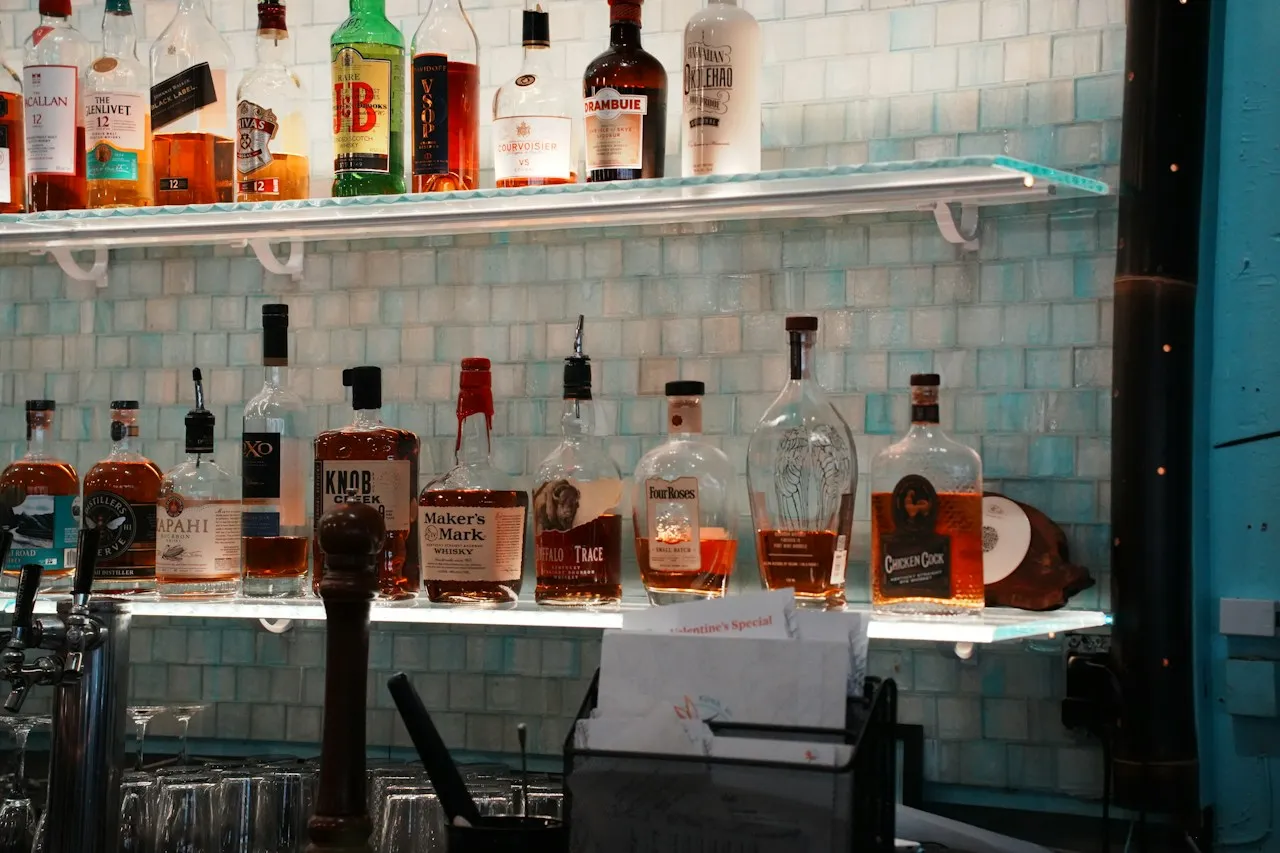 Bottiglie di superalcolici su due mensole di vetro illuminate di un bar, davanti a una parete di piastrelle turchese chiaro