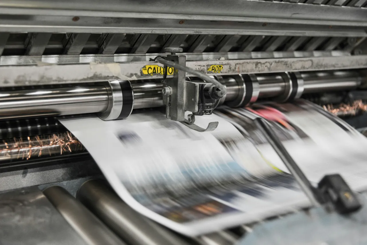 Wstęga papieru z zadrukowanymi stronami gazety przesuwająca się między stalowymi walcami maszyny drukarskiej