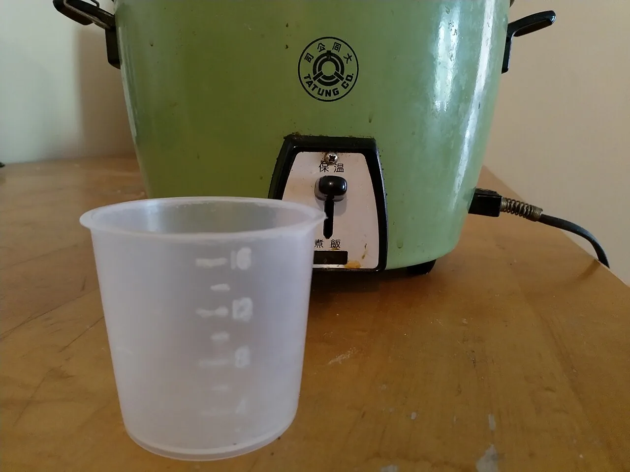 A plastic 180 millilitre rice measuring cup standing in front of a rice cooker
