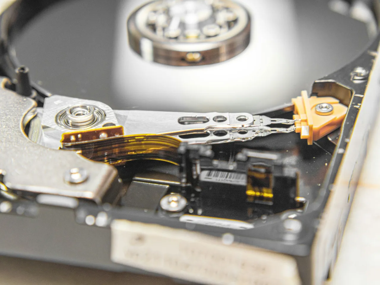 Inside an open hard drive: a shiny platter, with an arm holding the head and a ribbon cable above it