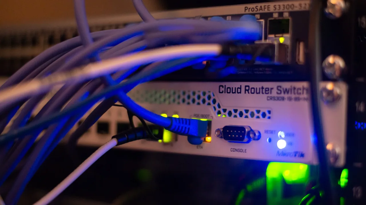 Przełącznik MikroTik Cloud Router Switch CRS309-1G-8S+IN z niebieskim przewodem w porcie ETH, nad nim urządzenie ProSAFE