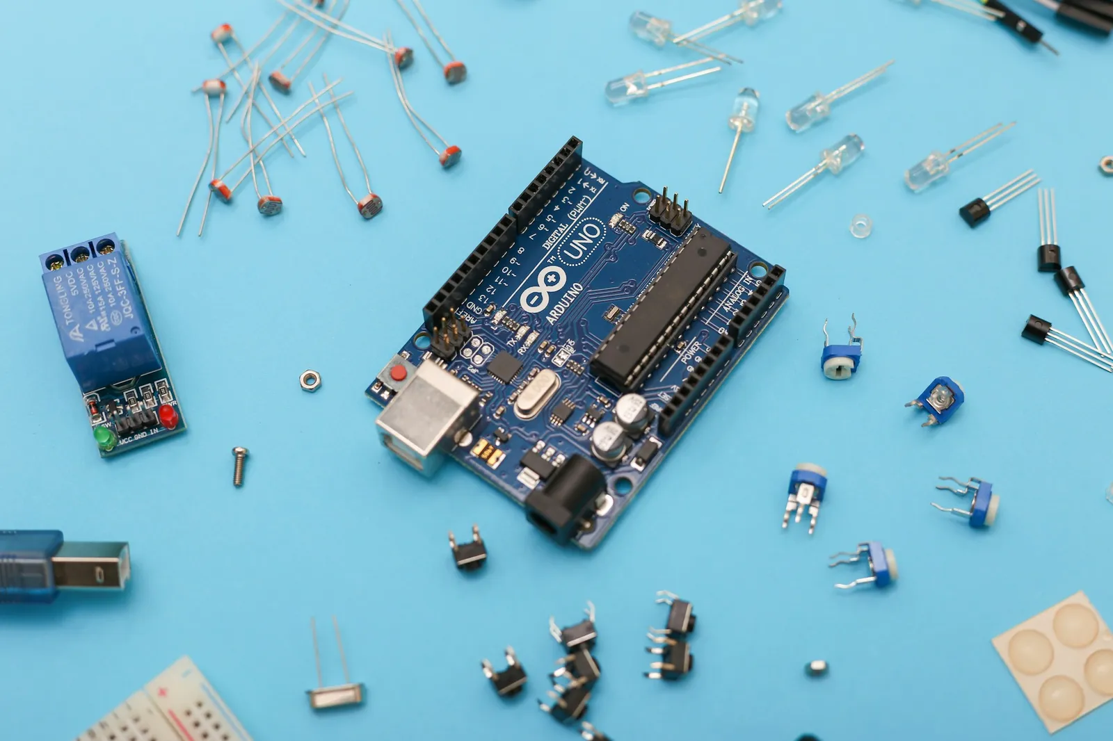 Płytka Arduino Uno z długim czarnym układem scalonym, a wokół niej przekaźnik, diody LED, fotorezystory i potencjometry na niebieskim tle