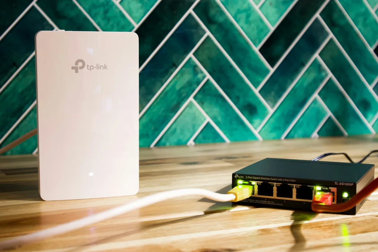 TP-Link TL-SG1005P, czarny przełącznik z pięcioma gniazdami, z żółtym i czerwonym przewodem, obok biały punkt dostępowy TP-Link
