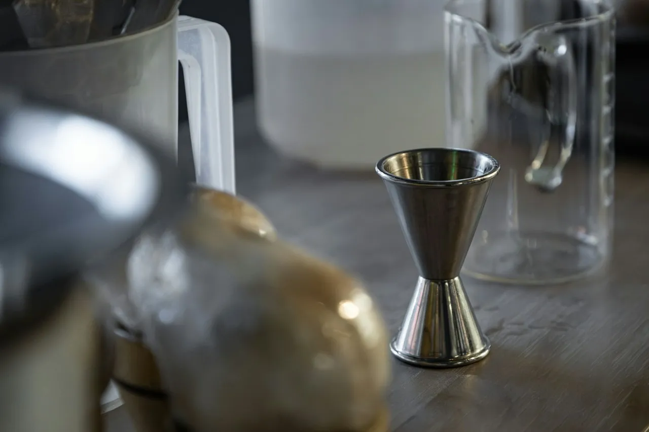 Un misurino da bar doppio in acciaio su un bancone di legno, con una caraffa di vetro dietro