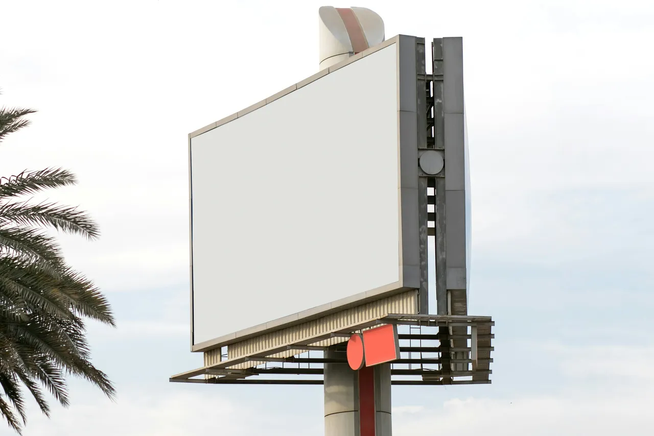 Painel publicitário branco e vazio num poste alto, junto a folhas de palmeira