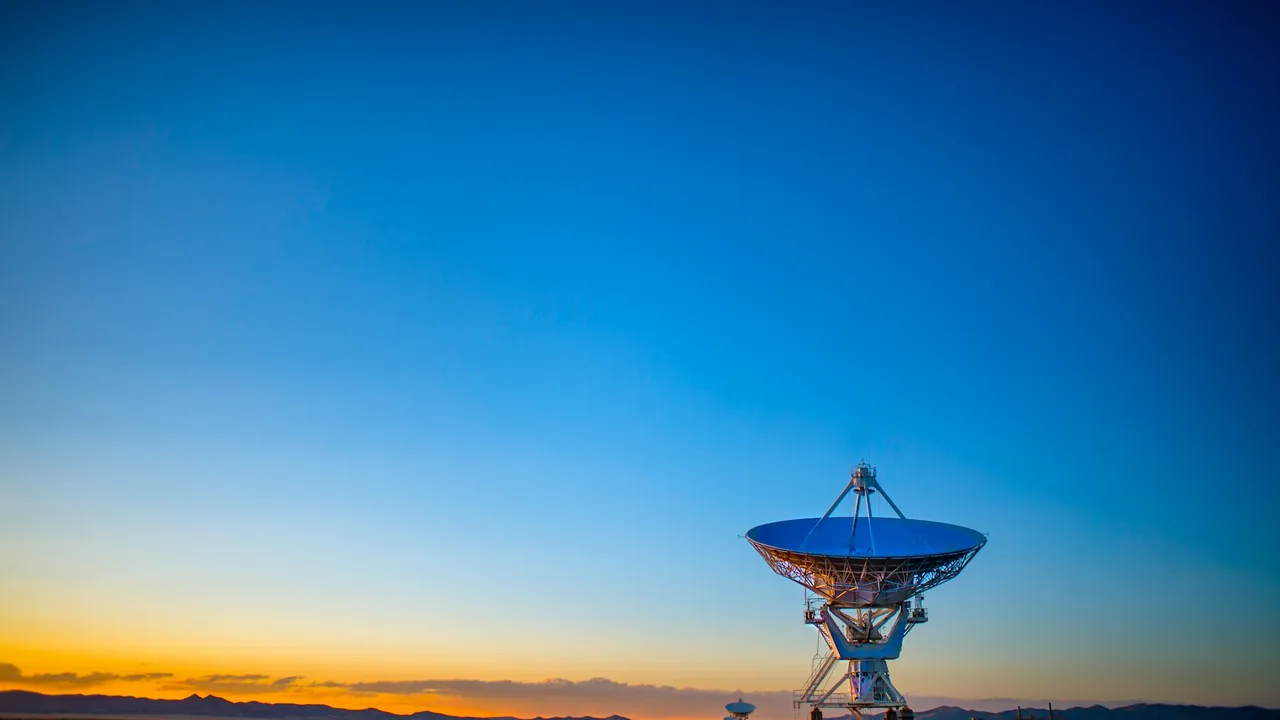 A radio telescope dish against a deep blue sky with an orange glow on the horizon, with a second, smaller antenna in the distance