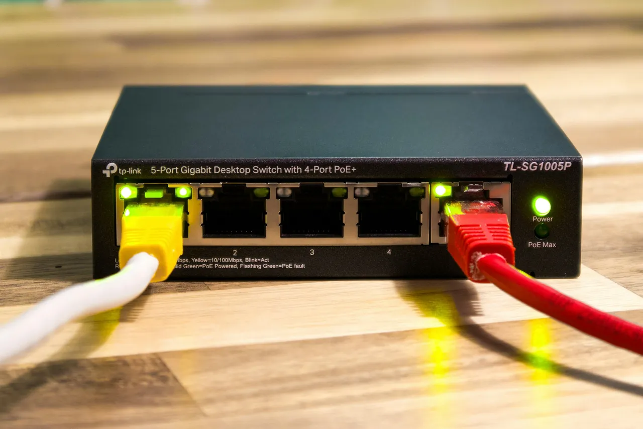 Czarne urządzenie sieciowe firmy TP-Link z pięcioma gniazdami i zielonymi lampkami na drewnianym blacie, do którego podłączono dwa przewody z żółtą i czerwoną wtyczką