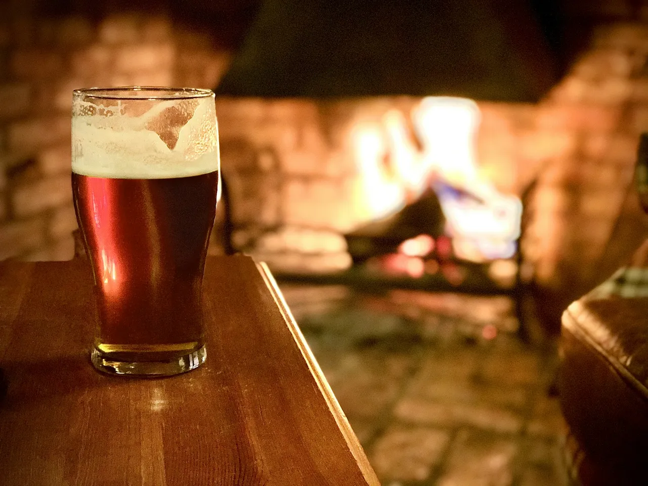 A glass of dark ale with a head of foam on a wooden table, with a fire burning in the background