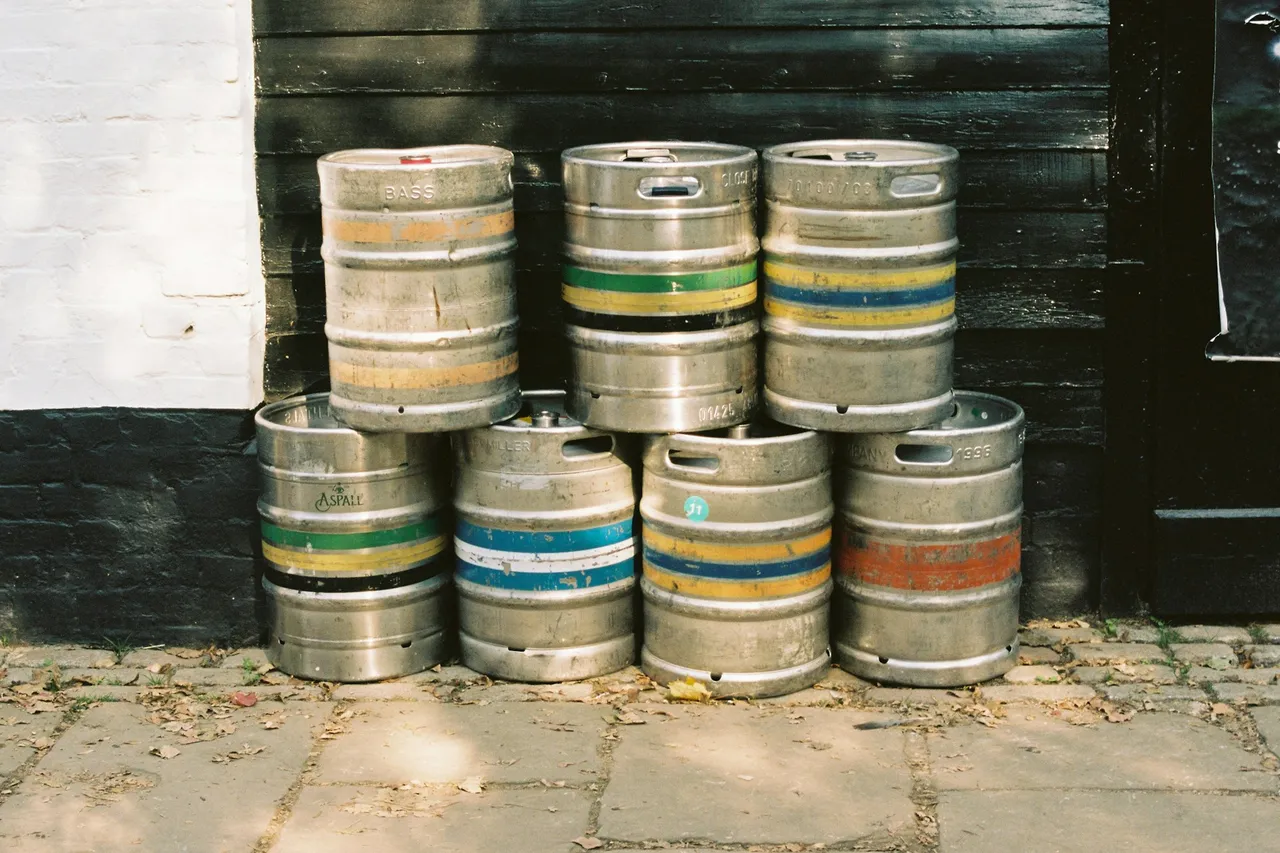 Seven steel beer kegs with coloured bands stacked in two rows against a black wooden wall